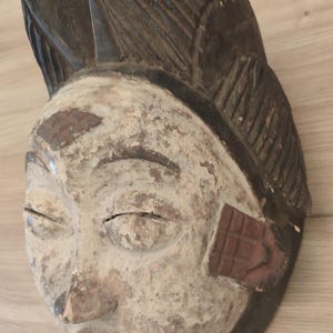 May include: A carved wooden mask featuring a white-painted face with dark brown lips, eyebrows, and hair. The hair is styled in a high, textured crest. The mask displays visible signs of wear, including chipped paint and a weathered, aged texture.