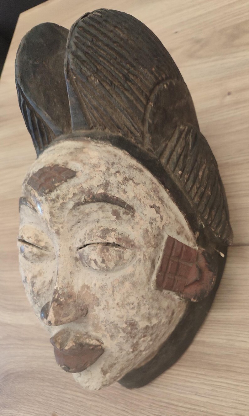 May include: A carved wooden mask featuring a white-painted face with dark brown lips, eyebrows, and hair. The hair is styled in a high, textured crest. The mask displays visible signs of wear, including chipped paint and a weathered, aged texture.