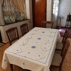 May include: A dining table set with a white tablecloth and matching napkins, featuring blue embroidered floral patterns. The table is surrounded by wooden chairs, creating an elegant dining arrangement. The room has a neutral colour palette.