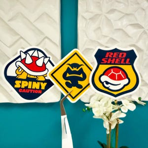 May include: Three colorful stickers featuring characters and symbols from a video game. One sticker reads "SPINY CAUTION" with a turtle shell design. Another is a yellow diamond with a black silhouette. The third is a red shell design with the words "RED SHELL".
