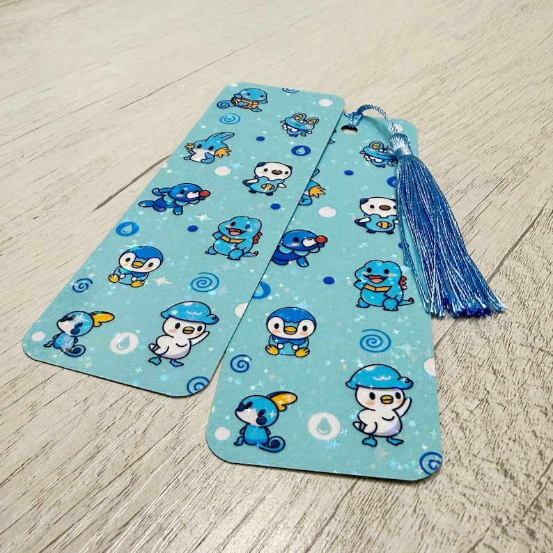 Puede incluir: Dos marcap&aacute;ginas de color azul claro con un patr&oacute;n repetido de personajes de dibujos animados y estrellas blancas. Un marcap&aacute;ginas tiene una borla azul. Los personajes son en tonos de azul, blanco y amarillo. Los marcap&aacute;ginas est&aacute;n sobre una superficie de madera clara.
