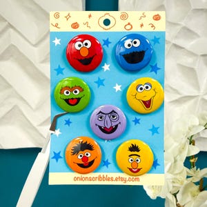 May include: A set of seven circular pin-back buttons featuring colorful Sesame Street character faces arranged on a light blue backing card with white star patterns and the text onionscribbles.etsy.com printed at the bottom of the display card.