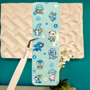 Puede incluir: Un marcap&aacute;ginas azul claro con varios personajes de dibujos animados. El marcap&aacute;ginas tiene un fondo azul claro con dise&ntilde;os de gotas de agua y remolinos azules. Los personajes de dibujos animados son en tonos de azul, blanco y amarillo.