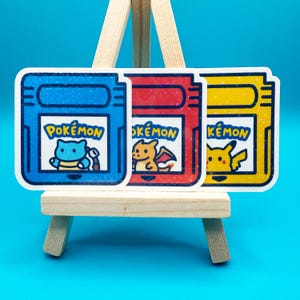 May include: Three colorful stickers featuring Pokémon characters. Each sticker has a rectangular shape with a blue, red, and yellow background. The stickers display the word "Pokémon" and a cartoon character.