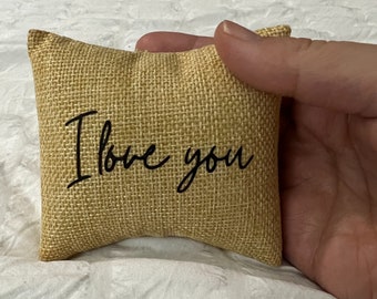 May include: A small, beige burlap pillow with the words "I love you" embroidered in black.