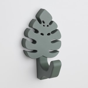 Puede incluir: Un gancho de pared con forma de hoja de monstera verde. El gancho está hecho de madera y tiene un diseño sencillo.
