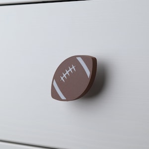 May include: A brown football-shaped cabinet knob with white stripes and stitching. The knob is mounted on a white cabinet door. This decorative knob adds a sporty touch to furniture.