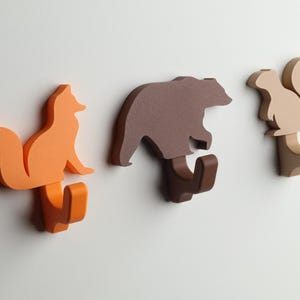 May include: Three wooden wall hooks shaped like a fox, bear, and squirrel. The fox is orange, the bear is brown, and the squirrel is beige.