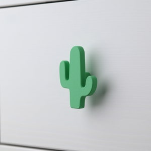 Puede incluir: Un tirador de cajón con forma de cactus verde para una cómoda blanca.