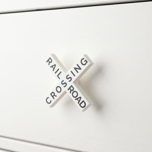 Può includere: Tiramaniglia per cassetto bianco a forma di segnale di passaggio a livello con la scritta "RAIL CROSSING" in nero.