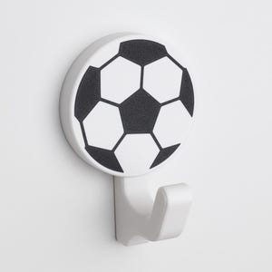 Pode incluir: Gancho de plástico branco com um design de bola de futebol. O gancho tem a forma de um J e tem uma base redonda com um gráfico de bola de futebol preto e branco.
