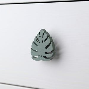 Op de afbeelding: Een witte lade met een groene, bladerige knop. Het blad is van hout en heeft een gedetailleerd ontwerp.