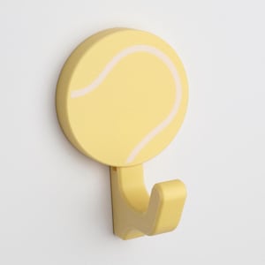 Puede incluir: Un gancho de pared amarillo con forma de pelota de tenis con una costura blanca.