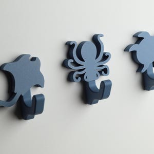 Puede incluir: Tres ganchos de pared azules con forma de pulpo, raya y tortuga. Los ganchos son de madera y tienen un diseño sencillo.