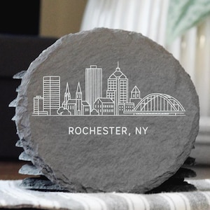 May include: A stack of gray slate coasters with a white line drawing of the Rochester, NY skyline. The coasters are round and have a natural, textured edge. The text "ROCHESTER, NY" is printed below the skyline.