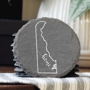 May include: A set of slate coasters featuring the outline of the state of Delaware with the text "home st" inscribed inside the state shape.