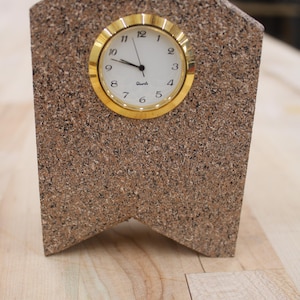 May include: A house-shaped desk clock with a speckled brown and tan granite-like finish. The clock features a circular gold-tone bezel surrounding a white face with black Arabic numerals and the word Quartz printed in the center of the dial.