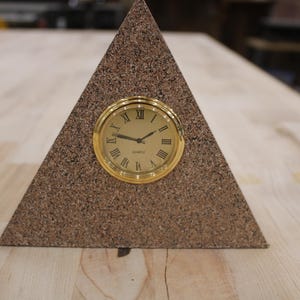 May include: A triangular desk clock with a textured brown speckled surface. The clock face is circular with a polished gold-tone bezel, black hands, and black Roman numerals. The word QUARTZ is printed in black at the bottom of the clock face.