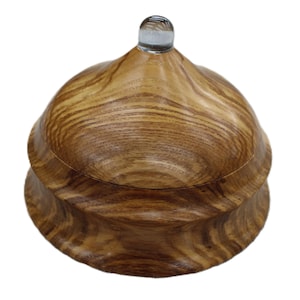 May include: A handcrafted wooden box with a lid. The box is made of light brown wood with a natural grain pattern. The lid has a clear glass knob on top. The box is round with a layered design.