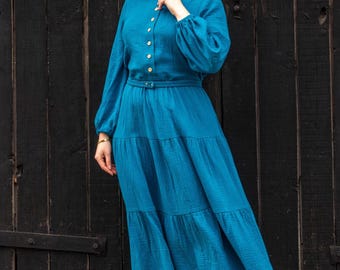 May include: A teal-colored dress with long sleeves and a tiered skirt. The dress features a button-down bodice and a belted waist. The fabric appears to be a lightweight, textured material. The dress is suitable for casual or semi-formal occasions.
