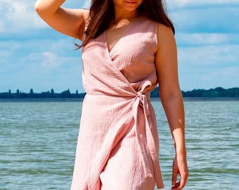 May include: A sleeveless, wrap-style dress in a soft, blush pink. The dress features a V-neckline and a tie closure at the waist. The fabric has a textured, crinkled appearance. The background shows a body of water and a blue sky with clouds.