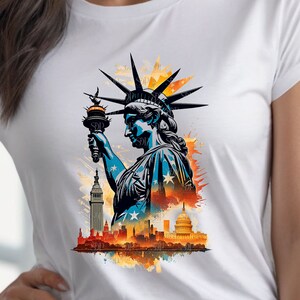 Op de afbeelding: Een wit T-shirt met een grafische print van het Vrijheidsbeeld dat een fakkel vasthoudt, met een stadsbeeld op de achtergrond. De grafiek is in aquarelstijl met spatten in oranje, geel en blauw.
