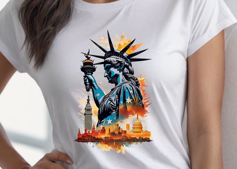 Op de afbeelding: Een wit T-shirt met een grafische print van het Vrijheidsbeeld dat een fakkel vasthoudt, met een stadsbeeld op de achtergrond. De grafiek is in aquarelstijl met spatten in oranje, geel en blauw.