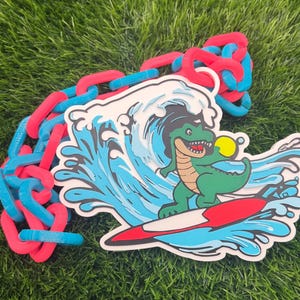 May include: A colorful image features a green dinosaur surfing a wave on a red surfboard, holding a yellow ball. The design is on a white background with blue and red chain links.
