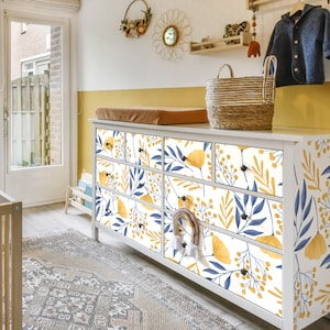 May include: White dresser with six drawers featuring a floral pattern in yellow and blue. The dresser has a white top and is against a yellow wall.