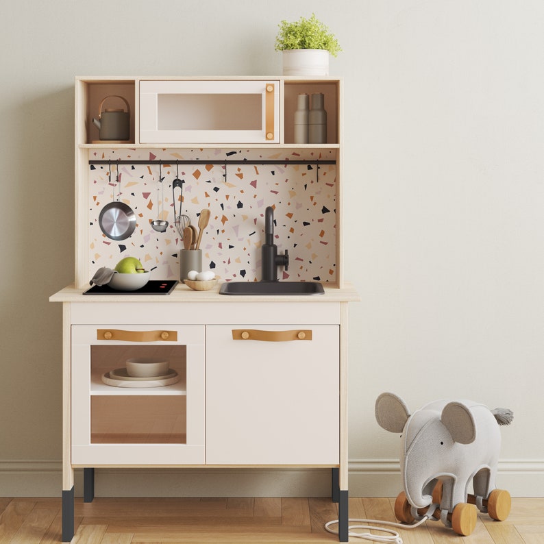 May include: A white wooden play kitchen with a black sink, a black stovetop, and a terrazzo-patterned backsplash. The kitchen has a cabinet with a glass door and a cabinet with a solid door. The kitchen has a shelf above the sink and a shelf above the stovetop. The kitchen has black legs. A gray elephant toy is in the foreground.