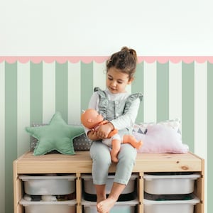 Op de afbeelding: Een kind zit op een houten opbergmeubel en houdt een pop vast. De kamer heeft een gestreepte muur in groen en wit, met een roze geschulpte rand. Een stervormig kussen en ander speelgoed zijn zichtbaar.