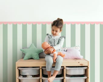 May include: A young child sits on a wooden storage unit, holding a doll. The room features a striped wall in shades of green and white, with a pink scalloped border. A star-shaped pillow and other toys are visible.