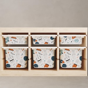 May include: Six white storage bins with lids, each featuring a repeating pattern of rockets, planets, and stars in blue, orange, and black on a cream background. The bins are arranged in a wooden shelf unit.