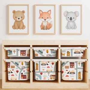 May include: A set of three framed animal prints featuring a bear, fox, and koala, above six white storage bins. The bins have a road and town design with cars, trees, and houses. The storage unit is made of light wood.