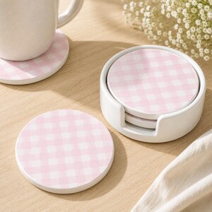 May include: A set of round ceramic coasters featuring a light pink and white gingham check pattern. The set includes a white ceramic holder for storage, with one coaster placed outside the holder and another positioned under a white ceramic mug.