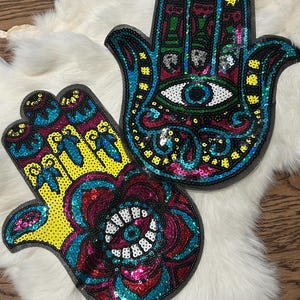 May include: Two sequined Hamsa hand patches. One is yellow, pink, and blue, with an eye design. The other is blue, pink, and black, also with an eye design. Both are on a white, furry surface.