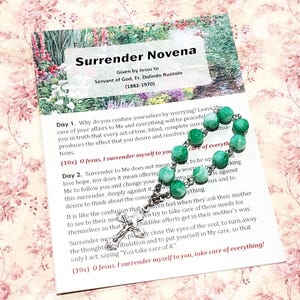 May include: A green and white rosary with a silver chain and a crucifix. The rosary beads are made of glass and have a marbled effect. The card in the background has the text "Surrender Novena" and a prayer for surrender to Jesus.