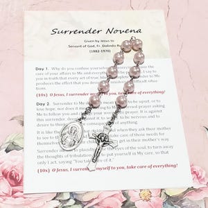 May include: A rosary with pale pink beads and a silver crucifix and medal, resting on a card with the text "Surrender Novena". The card has printed text about faith and prayer.