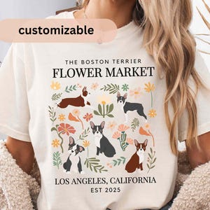 May include: A white t-shirt with a graphic design featuring Boston Terriers and flowers. The text "The Boston Terrier Flower Market" is printed on the shirt, along with "Los Angeles, California Est 2025".