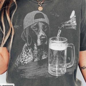 May include: A charcoal gray t-shirt featuring a graphic print of a spotted dog wearing a baseball cap, positioned next to a glass mug being filled with a liquid from a bottle. The shirt has a vintage-style wash and a crew neckline.