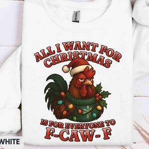 May include: White sweatshirt with a humorous Christmas graphic. The design features a grumpy rooster wearing a Santa hat and a sweater, surrounded by Christmas lights and holly. Text reads "ALL I WANT FOR CHRISTMAS IS FOR EVERYONE TO F-CAW-F."