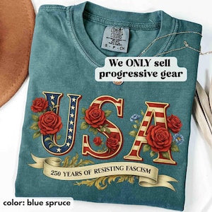 Op de afbeelding: Blauwspar T-shirt met een patriottisch "USA" ontwerp met rode rozen en een banner met de tekst "250 YEARS OF RESISTING FASCISM". Het shirt heeft ook de tekst "We ONLY sell progressive gear."