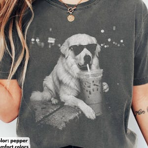 May include: Pepper-colored Comfort Colors t-shirt featuring a graphic of a dog wearing sunglasses and drinking from a clear iced coffee cup with a straw. The dog is sitting on a stone surface. The background has blurred lights.