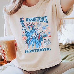 May include: Cream-colored t-shirt featuring a graphic of the Statue of Liberty in red and blue, with the words "RESISTANCE IS PATRIOTIC" in blue. The design includes fireworks and stars.