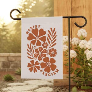 May include: A white garden flag with the words "RESIST FASCISM" in a rust-colored font. The flag features a floral design in the same rust color. The flag is displayed on a black metal stand in a garden setting.