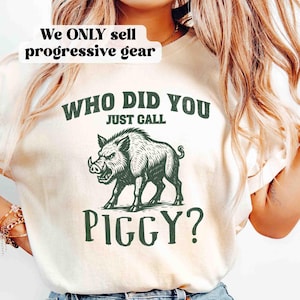 Op de afbeelding: Cr&egrave;mekleurig T-shirt met een groene illustratie van een wild zwijn en de tekst "WHO DID YOU JUST CALL PIGGY?". Het shirt heeft ook de tekst "We ONLY sell progressive gear" bovenaan.