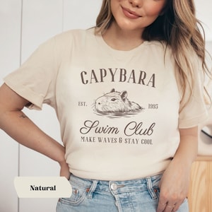 May include: A beige t-shirt with a brown graphic of a capybara swimming. The text "Capybara Swim Club Est. 1995 Make Waves & Stay Cool" is printed on the shirt.