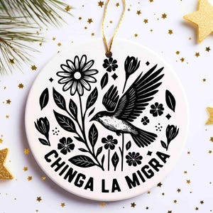 May include: White ceramic ornament with a black and white floral and bird design. The ornament features a daisy, leaves, and a bird in flight. The text "CHINGA LA MIGRA" is printed in black around the bottom edge. A gold string is attached for hanging.