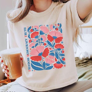 May include: Ivory-colored t-shirt featuring a floral design with red and pink flowers, blue leaves, and the text "FREEDOM DEMOCRACY TRUTH." The shirt is a neutral color and has a relaxed fit.