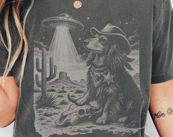 May include: Pepper-colored t-shirt with a graphic of a dog in a cowboy hat and bandana, with a sheriff's badge, under a UFO beam in a desert landscape. A cow skull and cactus are also in the image. The text on the shirt says "comfort colors".
